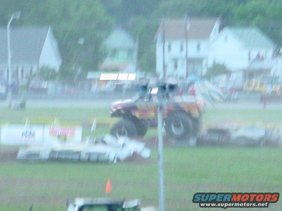Monster truck with orange and yellow graphics, large tires, and dual exhaust pipes, angled to the front