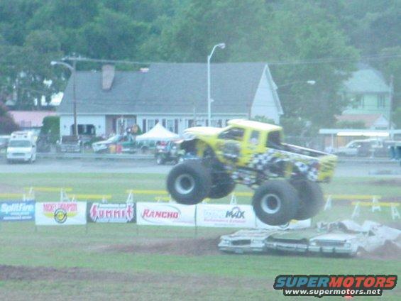 Yellow monster truck mid-air over crushed cars, checkered flag pattern on rear
