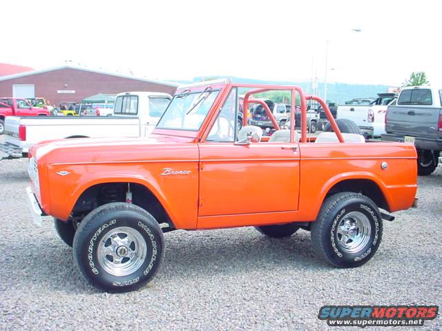 Orange 1979 Ford Bronco with a roll cage, parked on gravel with other trucks in the background