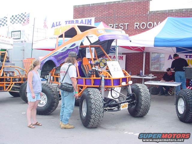 Orange and purple custom off-road vehicle with large tires and roll cage, two people standing nearby