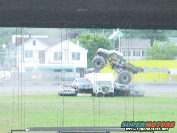 Monster truck on top of crushed cars, with houses in background