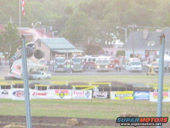 Monster truck flipped on its side with large tires visible, at an outdoor event with other trucks and buildings