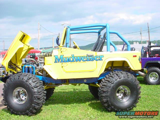 Yellow off-road vehicle with blue roll cage and 'Mudweiser' text. Hood is open, showing engine components. A purple vehicle is parked behind it