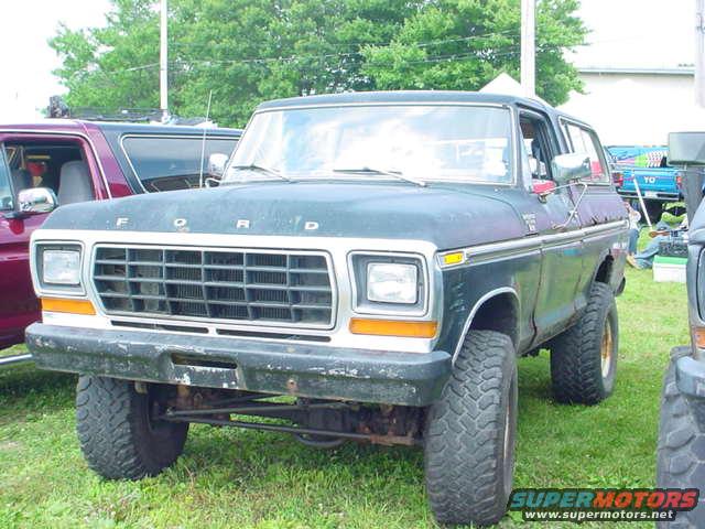 Front view of a dark colored Ford Bronco with a large grille and off-road tires. Another vehicle is partially visible to the left