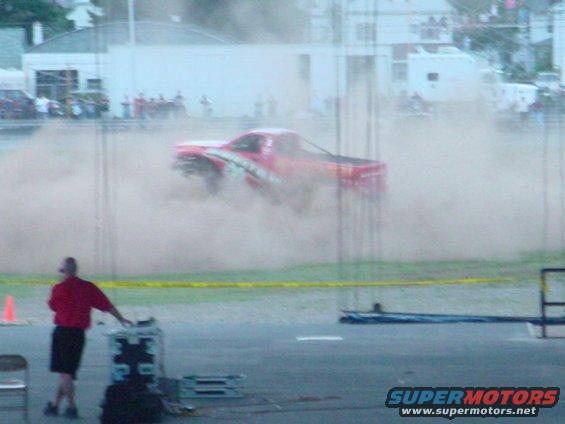 Red pickup truck kicking up dust during a burnout, with a person in a red shirt watching