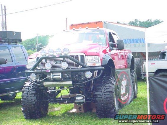 Red lifted Ford F-series truck with custom bumper, winch, and multiple lights. Another vehicle is partially visible to the left