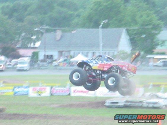 Monster truck with red, yellow, and blue paint jumping over crushed cars