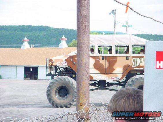 Monster truck with a tan and brown camouflage pattern and a white bus-like top, people inside. Text 'SERGE' visible on the side
