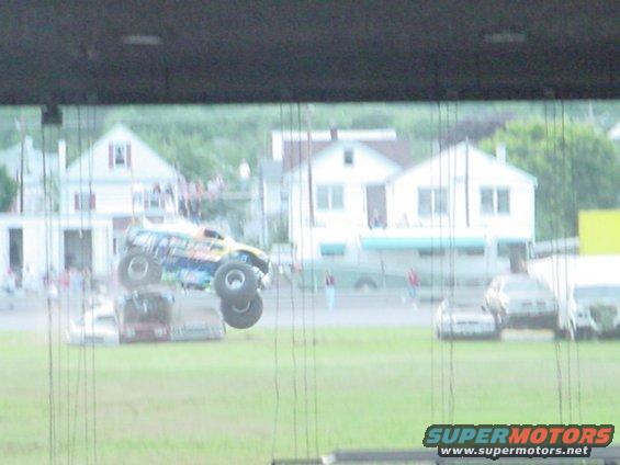 Monster truck airborne over crushed cars, with houses and spectators in background