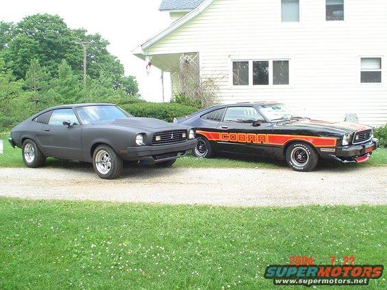 Two matte black and black/orange 1978 Ford Mustang IIs parked on a gravel driveway in front of a house