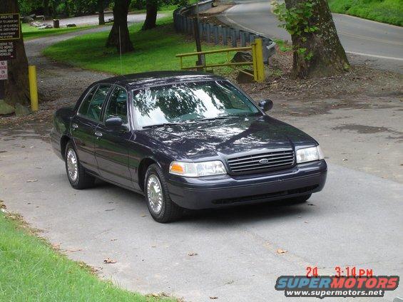 Dark blue sedan parked on a paved driveway with trees and a guardrail in the background. Watermark reads SUPERMOTORS.NET