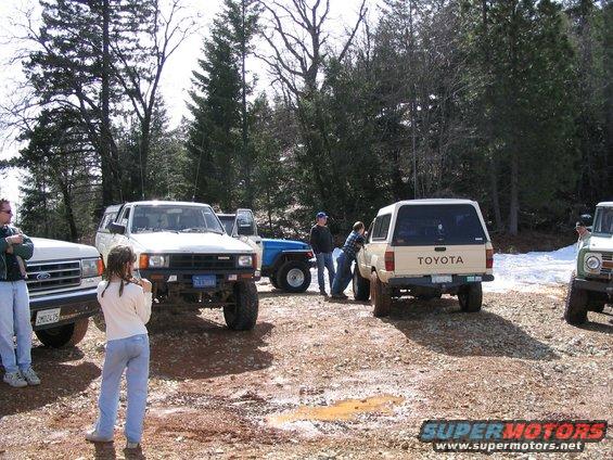 Several off-road vehicles parked on a dirt lot with trees in the background. A white Toyota SUV is visible