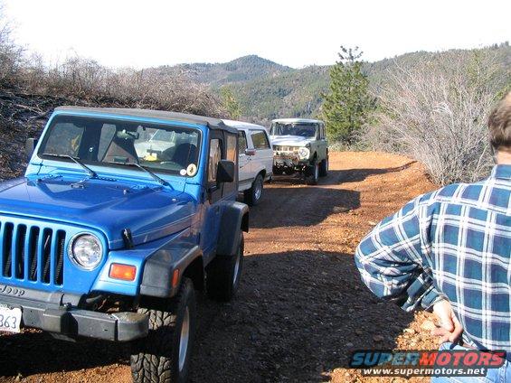 Blue Jeep Wrangler and white Ford Bronco on a dirt road with hills in background