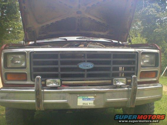 Front view of a 1980s Ford Bronco grille with headlights and bumper. Hood is open