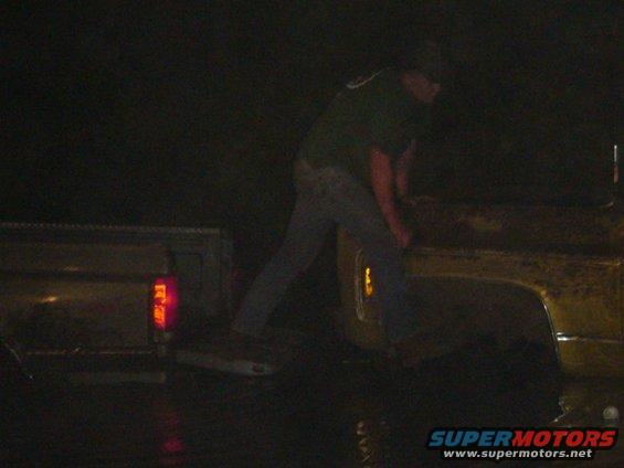 Rear view of a truck bed with a person loading it at night. Water on the ground reflects the taillight