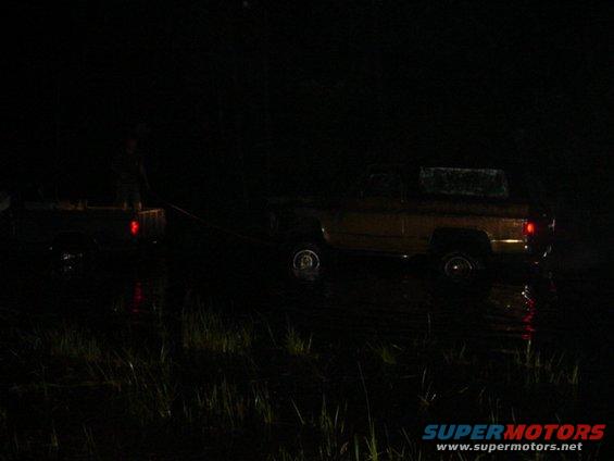 Two vehicles are partially submerged in water at night, with one appearing to be towed