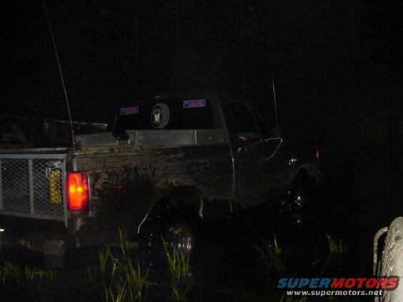 Mud-covered Ford truck in water at night, rear view. Diamond plate toolbox on bed, two antennas, and a mesh tailgate