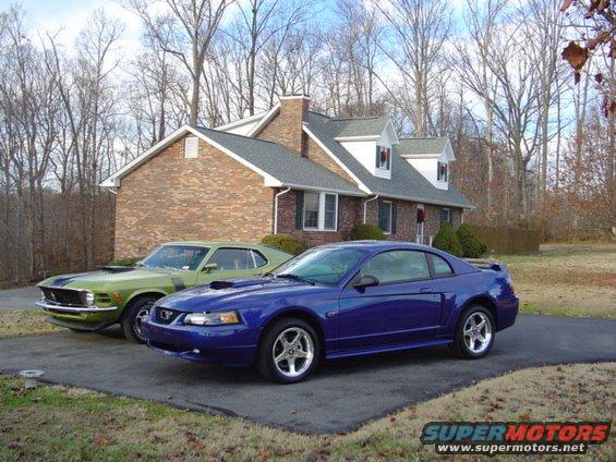 1970 Ford Mustang in green with black stripes and a blue Ford Mustang parked in front of a house