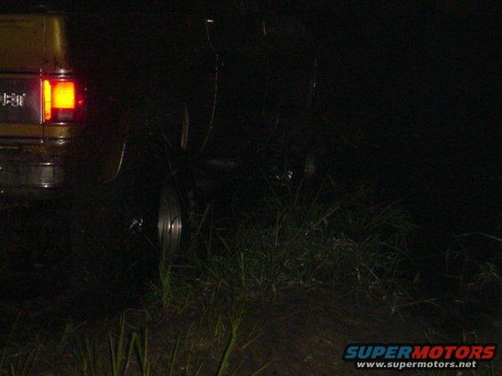 Yellow vehicle's illuminated rear tail light and tire are visible in the dark, partially obscured by grass