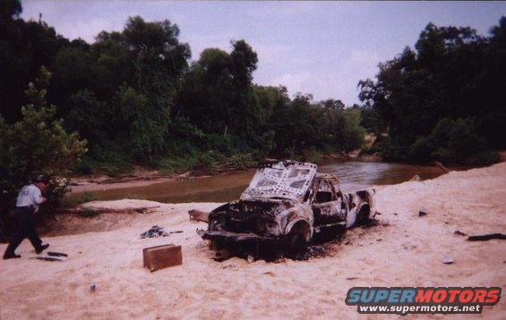 Burnt pickup truck on sandy riverbank, hood open, interior gutted, with a person in uniform standing nearby