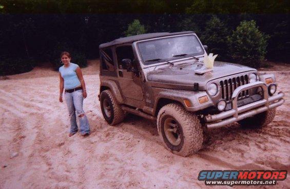 Brown Jeep Wrangler with a soft top, parked on sand with a woman standing nearby