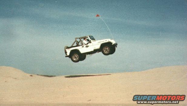 White Jeep Wrangler airborne over sand dunes with a red flag attached