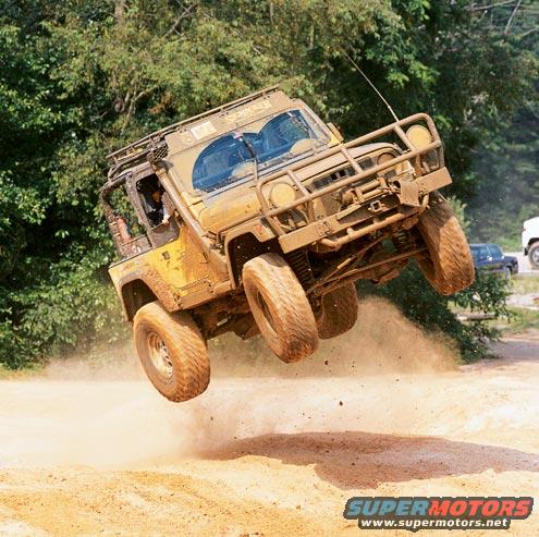 Mud-covered off-road vehicle airborne over dirt track, trees in background. Another vehicle visible in distance