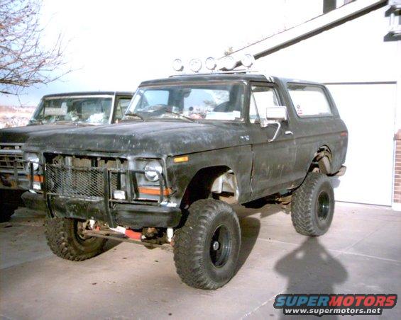 Black 1978 Ford Bronco with a brush guard and roof lights, parked in front of a garage