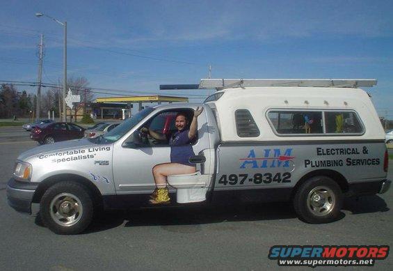 Ford F-Series truck with a person sitting on a toilet in the driver's seat, waving. Truck has "AIM Electrical & Plumbing Services" and phone number on the side