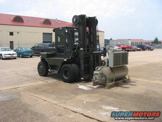Forklift with an air compressor attached, parked on concrete. Other cars are in the background