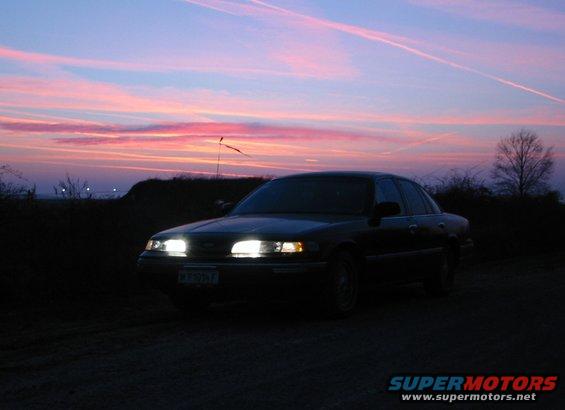 1992 Ford Crown Victoria parked on dirt road with headlights on, sunset sky