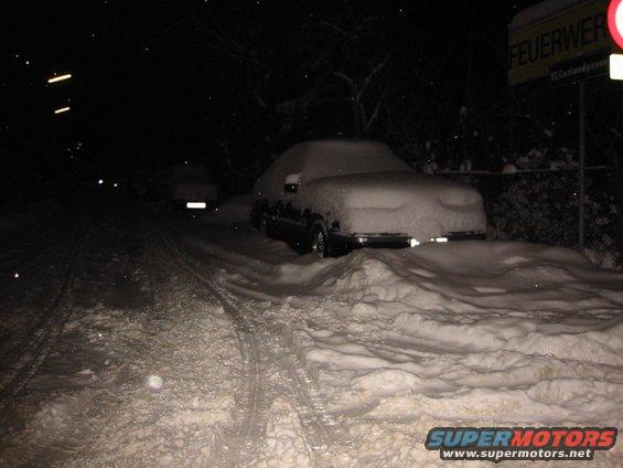 Two cars are buried in snow on a dark, snowy road. A sign reads FEUERWEHR