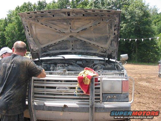 Front view of a muddy 1987 Ford F-Series truck with hood open, showing the engine bay. Two men are looking at the engine