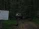1988 Ford Bronco - A tan off-road vehicle drives down a wooded trail past a sign warning of risk.