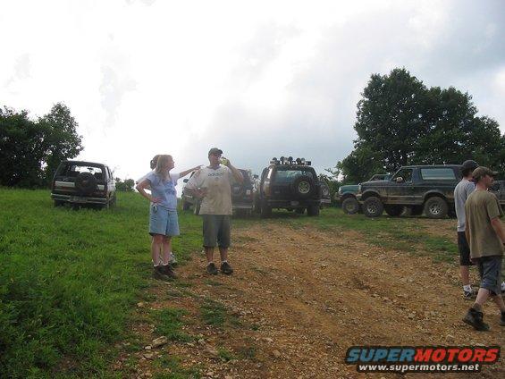 Several off-road vehicles parked on a dirt path with people standing around. One Bronco is prominent