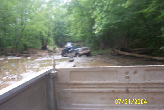 Two trucks are stuck in a muddy creek bed surrounded by trees. The truck in the foreground is a light-colored pickup bed