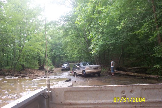 Two SUVs navigate a muddy forest stream, one in the foreground, another further back. A person stands on the bank