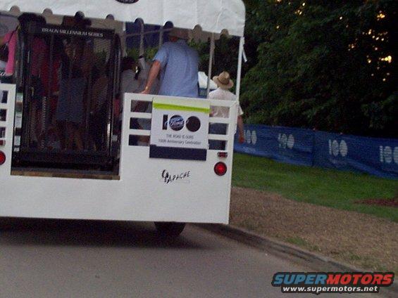 White shuttle bus with 'Ford 100' logo and 'The Road is Ours' text. 'Apache' is written on the back
