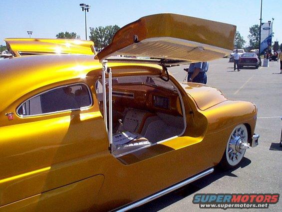 Customized classic car with gold paint, gullwing doors open, showing wood-paneled interior and white-wall tires