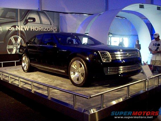 Dark blue concept car on display platform, front quarter view. Large chrome grille and distinctive headlights