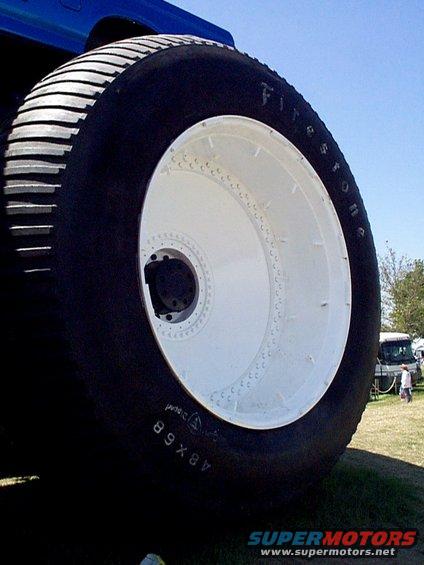 Close-up of a massive tire with 'Firestone' visible on the sidewall. A blue vehicle is partially visible above. Other vehicles and people are in the background
