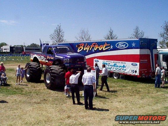 Purple Bigfoot monster truck parked next to its red, white, and blue trailer. Several people stand around the truck and trailer