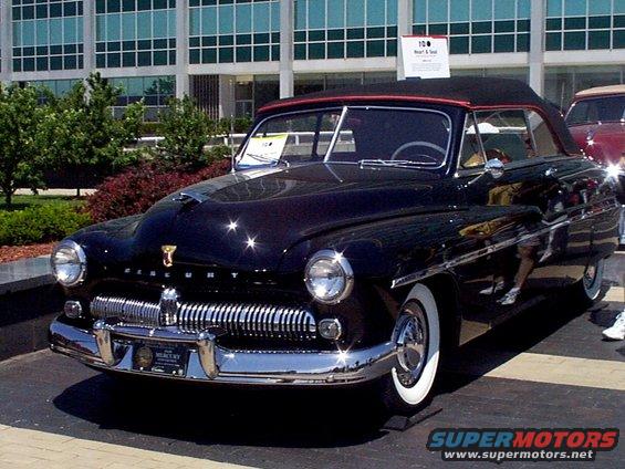 Black 1950 Mercury convertible with red top, parked outside a modern building. Another car is partially visible to the right