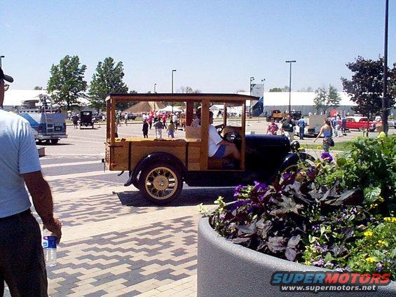 Wooden wagon with a driver, seen from the side. Other vehicles and people in background