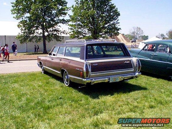 Rear view of a brown station wagon with wood paneling and a roof rack parked on grass. Another car is visible to the right