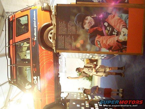Orange expedition vehicle with 'G4 Challenge' and 'The ultimate global adventure' text, parked next to display with kids