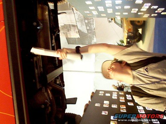 Person in a tan hat and t-shirt holds a rolled document near a red object and a table with many small photos