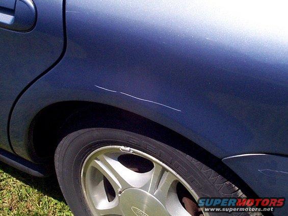 Dark blue car rear quarter panel with scratches above a silver wheel. Grass visible below