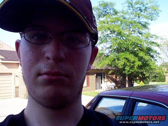Person wearing a baseball cap and glasses takes a selfie in front of a maroon car and a house