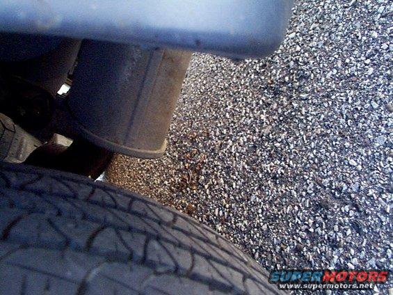 Close-up of a vehicle's exhaust pipe and tire on a gravel surface. Supermotors.net logo visible